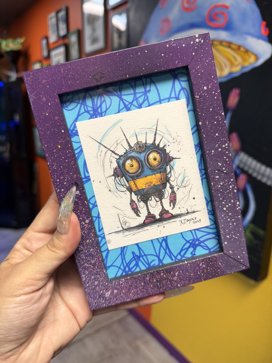 XZanthia's hand holding up a small purple-framed creature watercolor