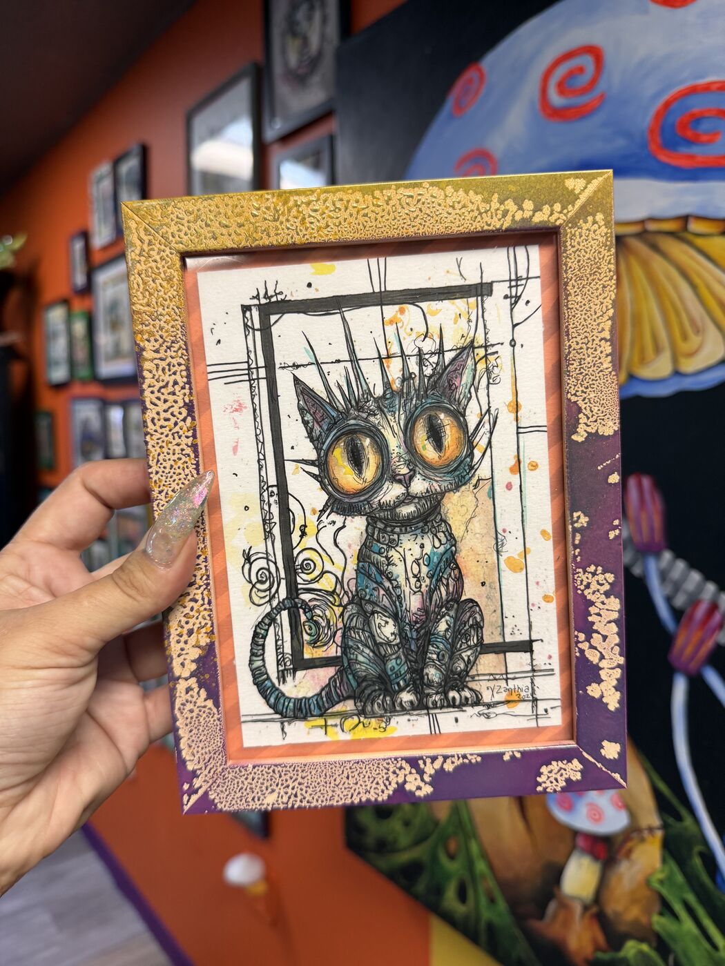 XZanthia holding up a framed creepy-cute cat watercolor in her studio